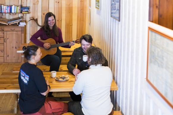 4 Personen sitzen zusammen bei Gebäck und Kaffee, eine Person davon spielt Gitarre